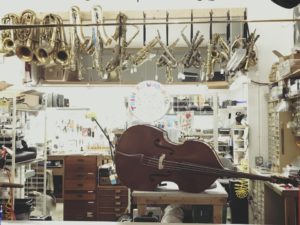 A string bass lies on its side on a cart in the middle of a repair shop with tall ceilings, with broken saxophones and brass instruments hanging from large hooks near the ceiling.